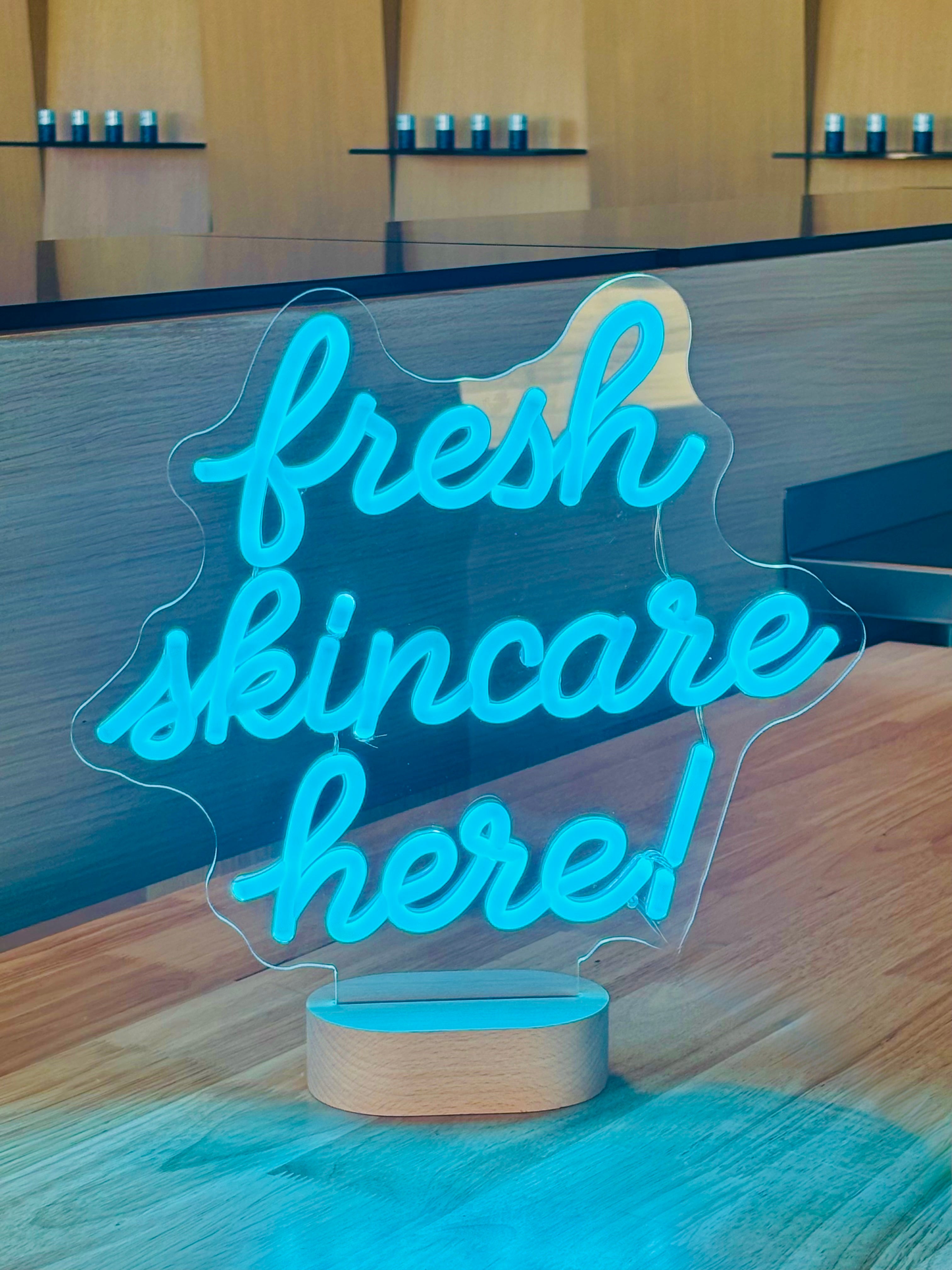 A neon-style 'fresh skincare here!' sign on a wood counter, with Skin at Peace products on shelves behind.
