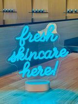 A neon-style 'fresh skincare here!' sign on a wood counter, with Skin at Peace products on shelves behind.